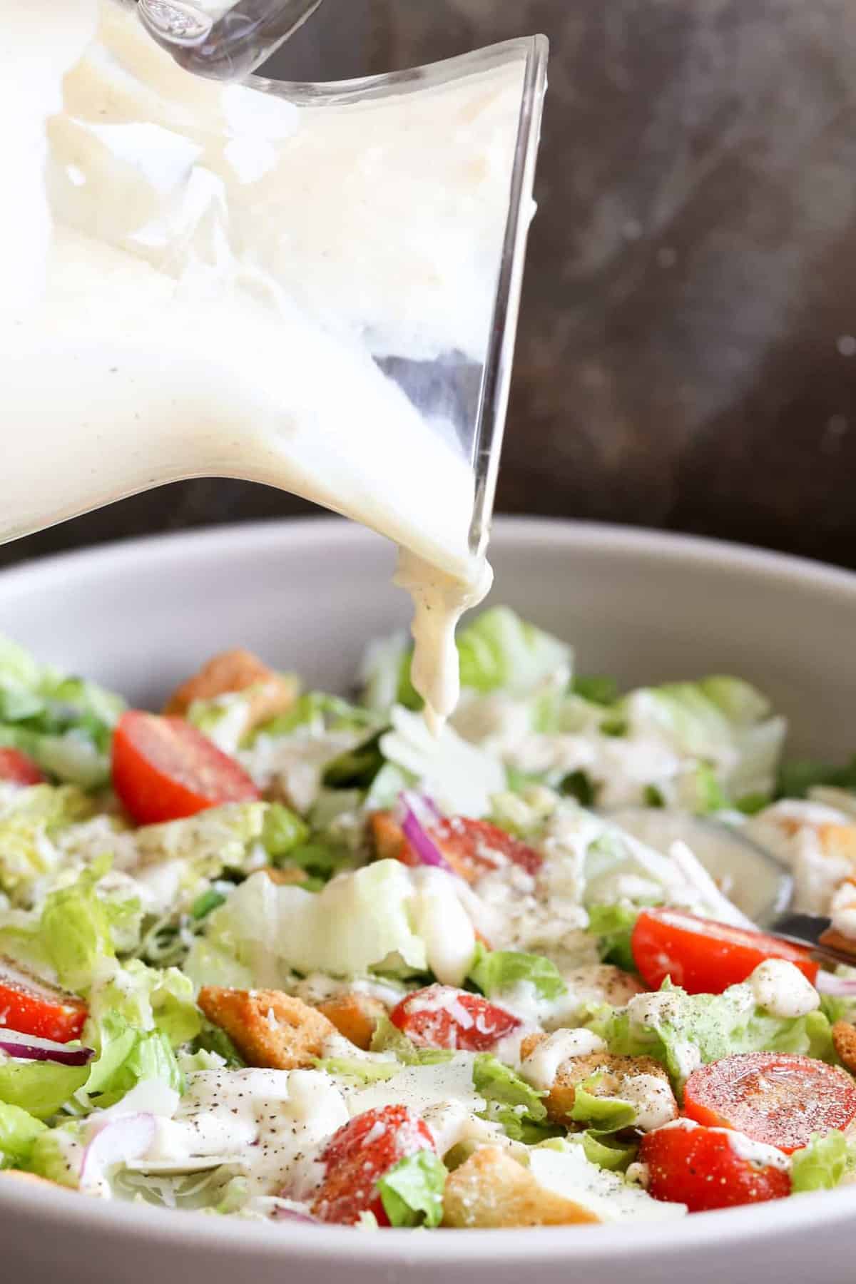 Caesar salad dressing pouring into salad bowl that is loaded with lettuce, tomatoes, croutons, and onions.