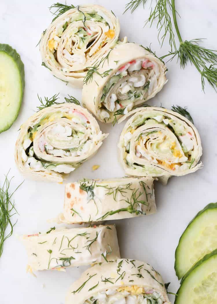 Tortilla and crab pinwheels on a board topped with fresh dill, next to cucumber slices and dill. 