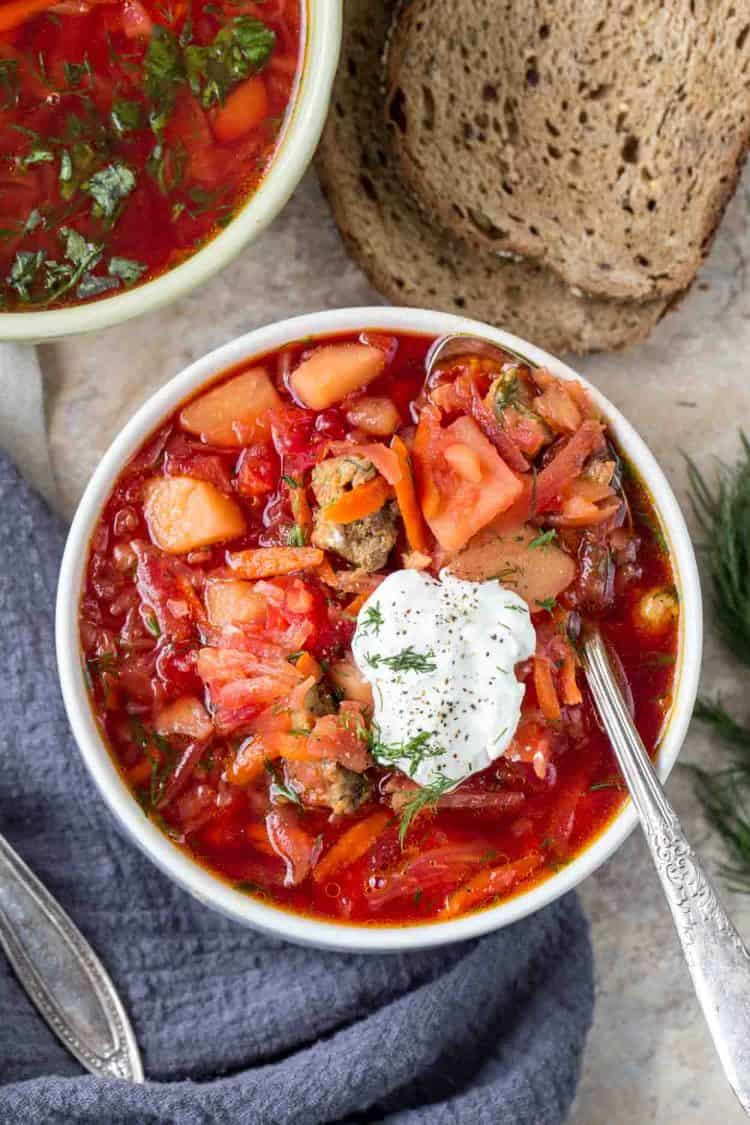 A bowl of Russian borscht with sour cream and fresh herbs with dill and bread slices next to the bowl.
