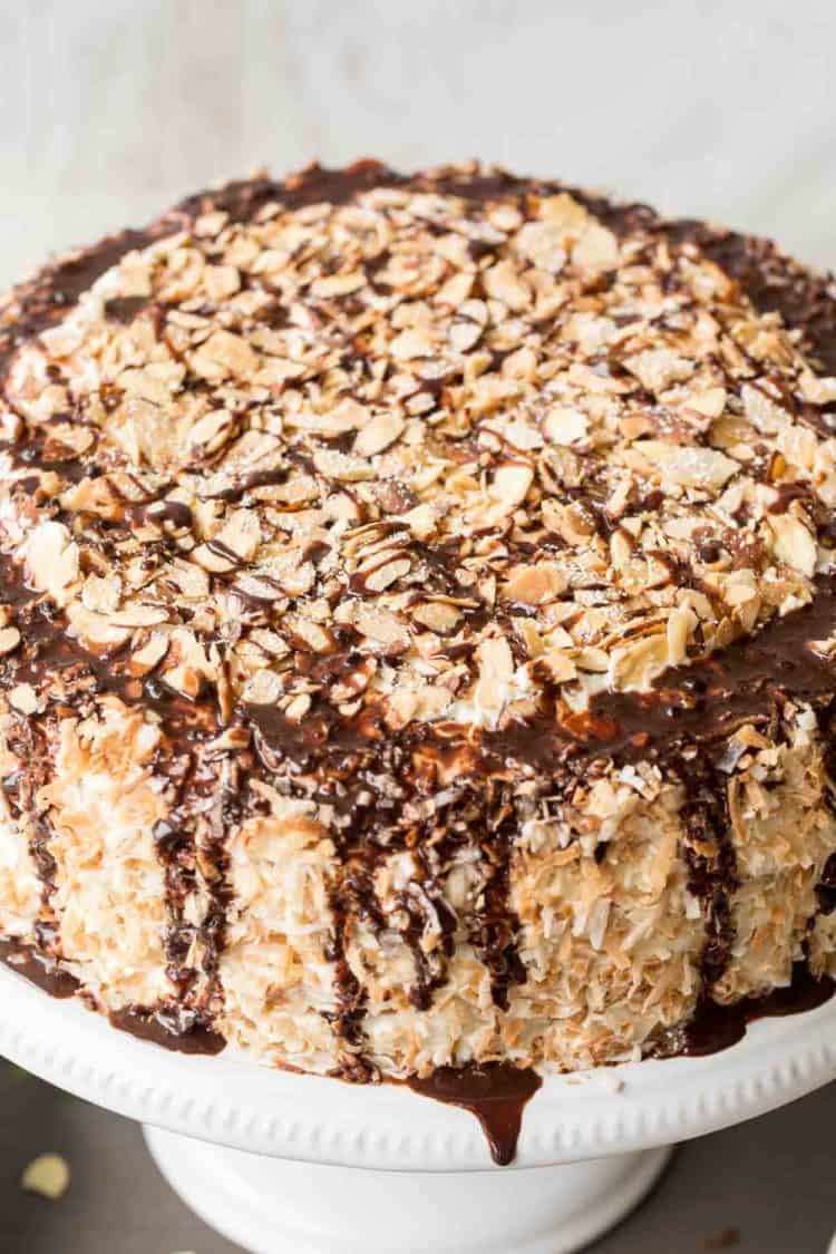Coconut cake on a platter topped with toasted coconut, almonds, and chocolate drizzle.