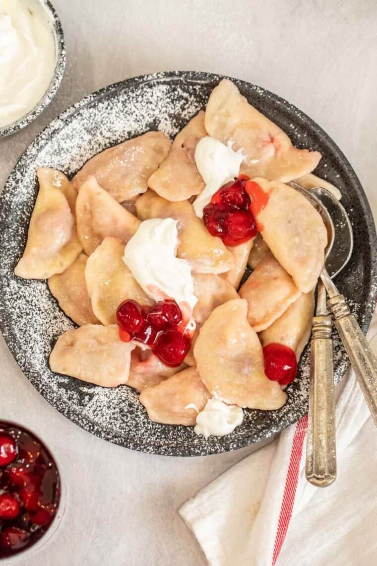 Vareniki on a black plate topped with cherries and sour cream. 