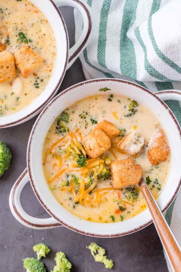 Two bowls full of broccoli cheese soup next to a white and green rag and broccoli florets. 