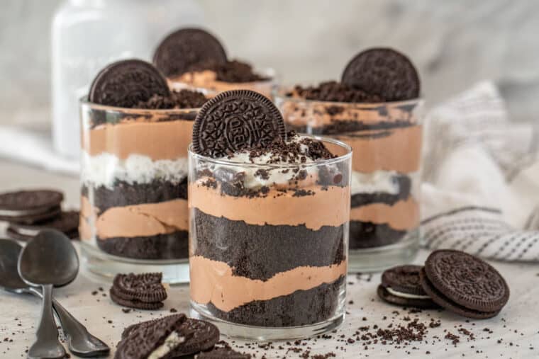 Oreo pudding in cups topped with Oroes next to black spoons and Oreo crumbs. 