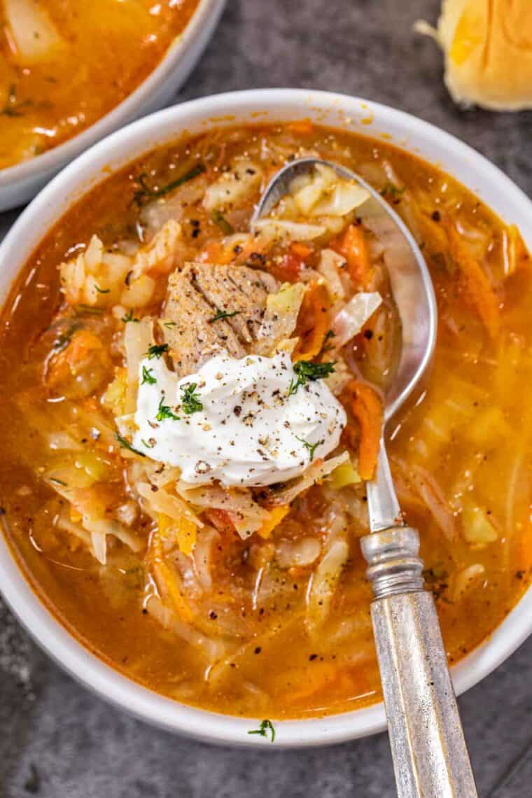 Beef and cabbage soup in a white bowl with a metal spoon topped with sour cream and fresh dill. 