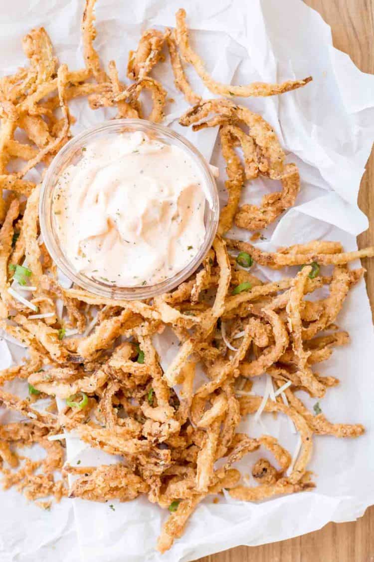 Fried onion straws next to a bowl of dipping sauce on white parchment paper.