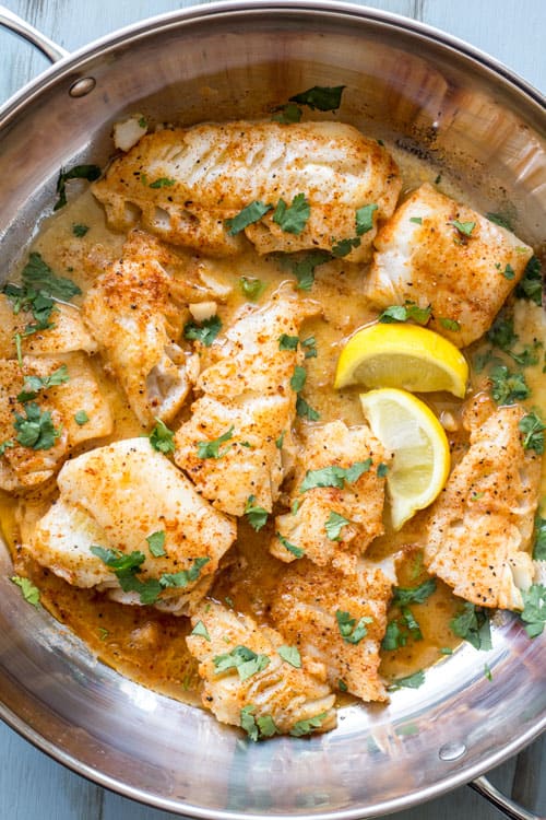 Cod fish in a pan with lemon wedges and herbs.