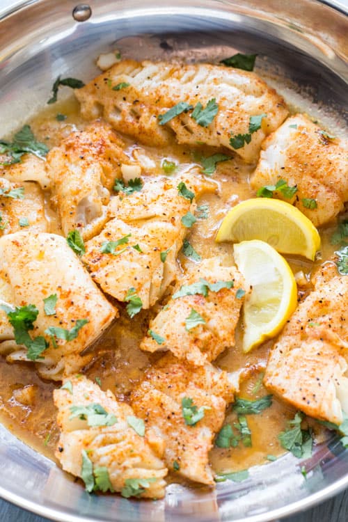 Cod fish fillets in a skillet in a butter sauce and topped with lemon wedges and herbs.