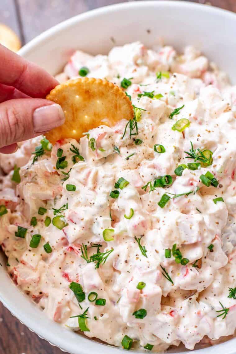 A white bowl with cold crab dip with a cracker being dipped into the dip. 