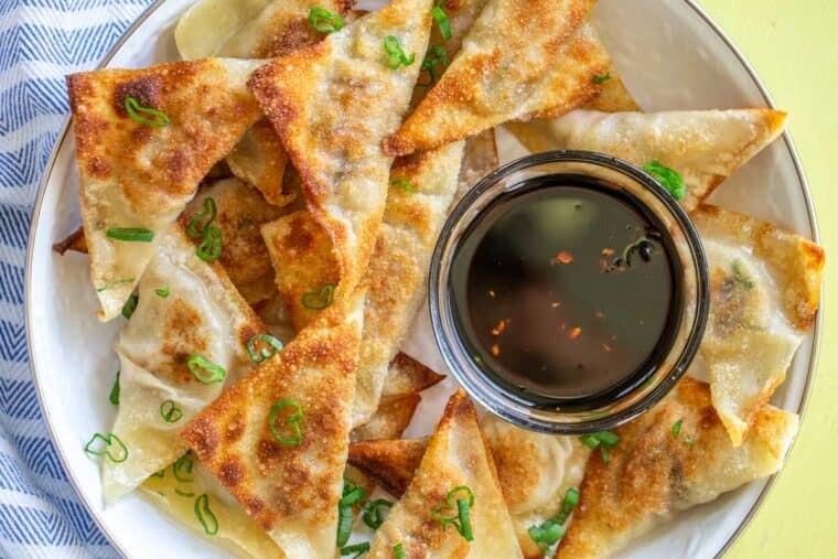 A white plate of pork potstickers topped with green onion next to a bowl of dipping sauce. 