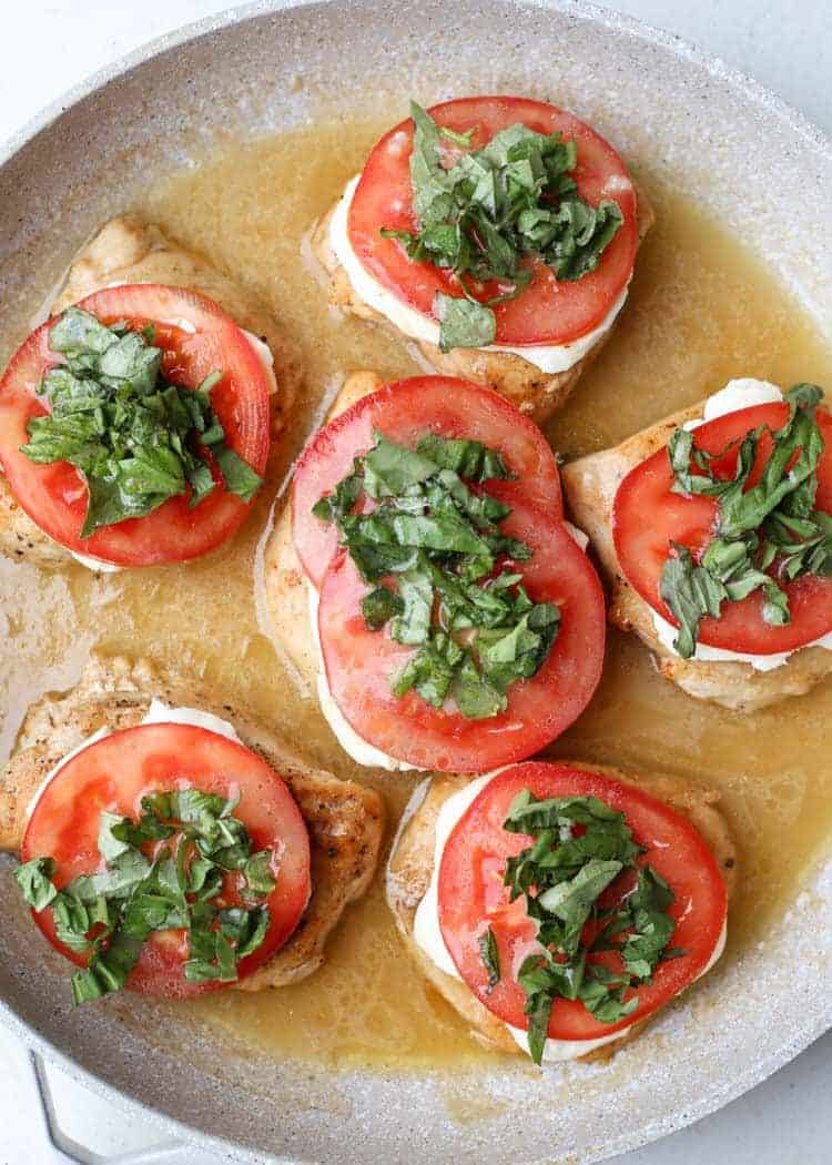 Chicken breast topped with cheese, tomato, basil in a white wine sauce in a skillet.