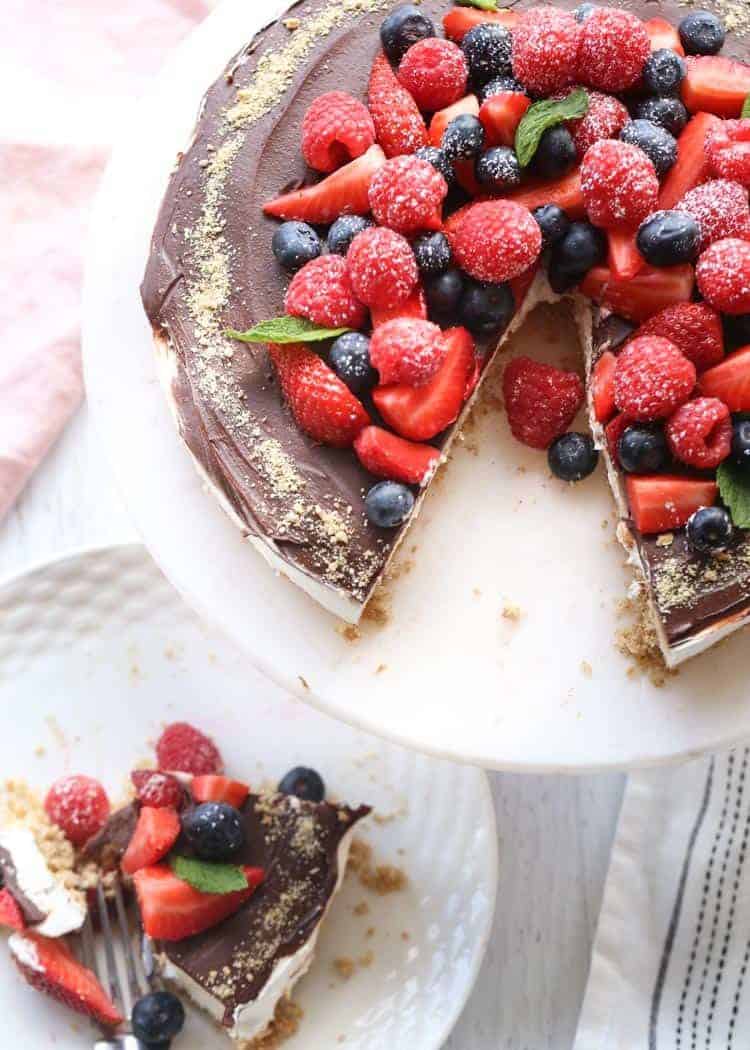 No bake cheesecake recipe with berries and a chocolate ganache on a platter, with a slice cut.
