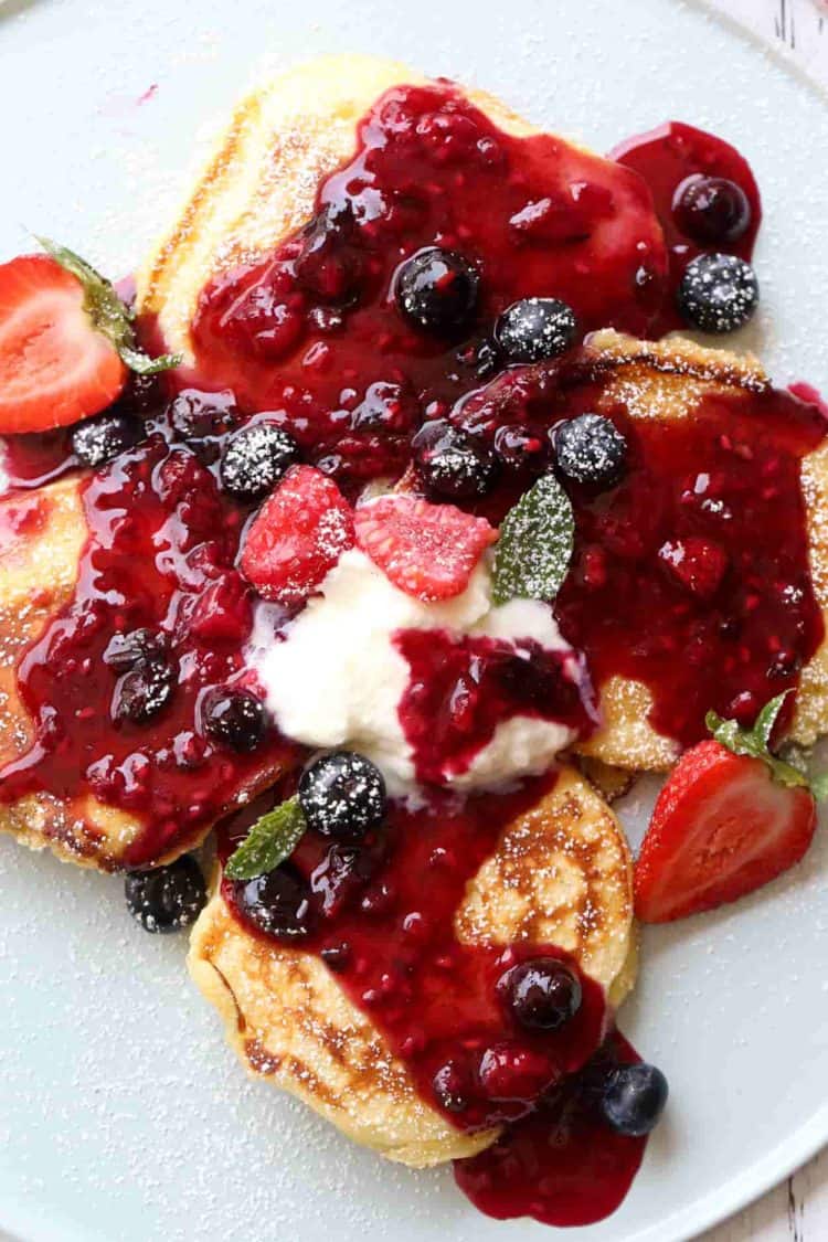 Four Ricotta Pancakes on a plate with berry syrup, fresh berries, and powdered sugar sprinkled on top.