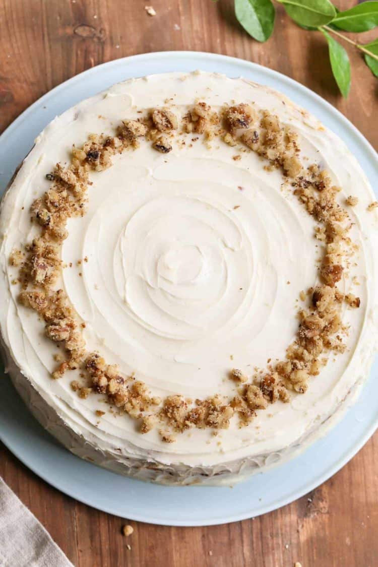 Carrot cake on a cake stand with cream cheese frosting and crushed walnuts on top.