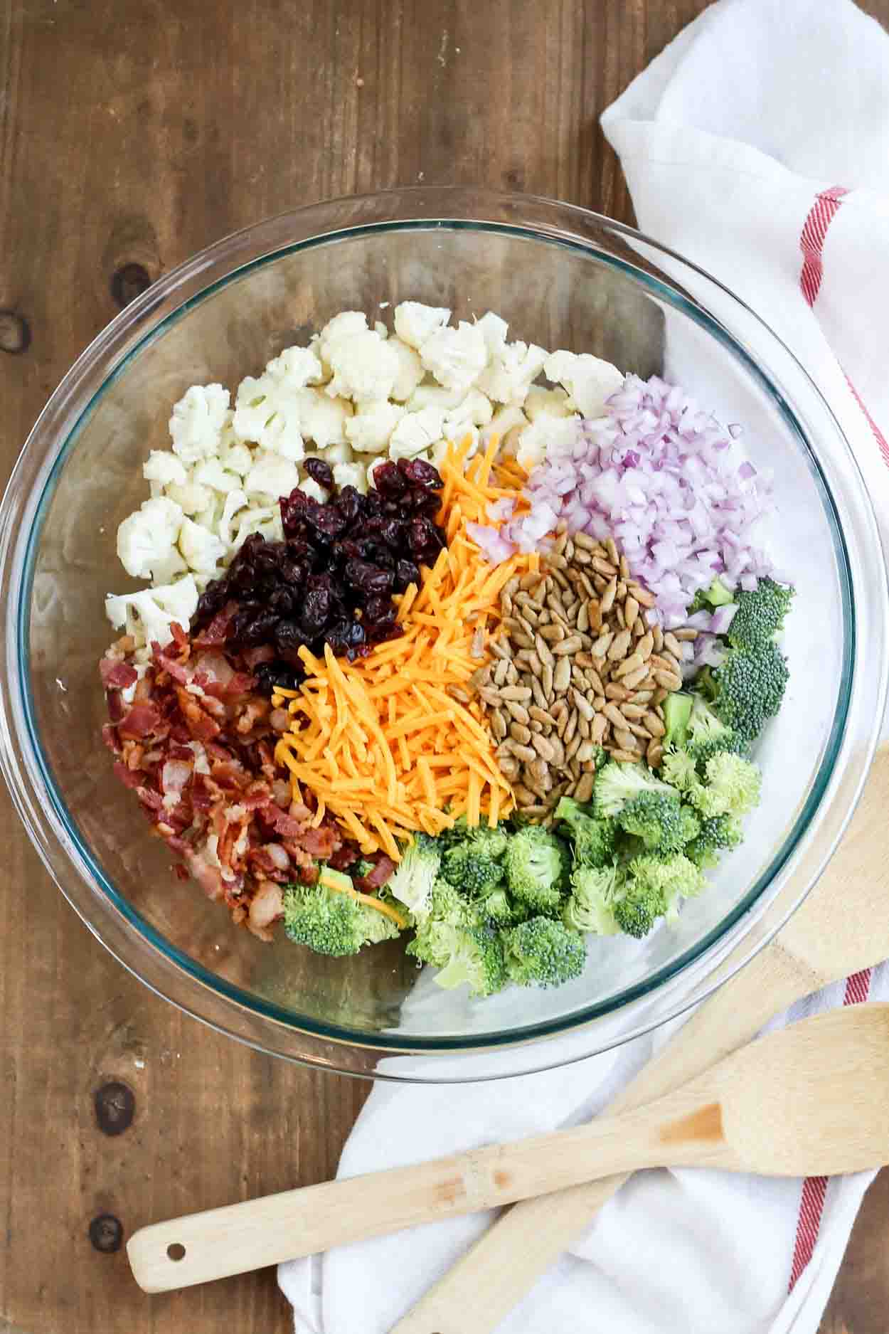 Simple cauliflower broccoli salad recipe in a bowl with cheese, onions, craisins, sunflower seeds, and bacon.