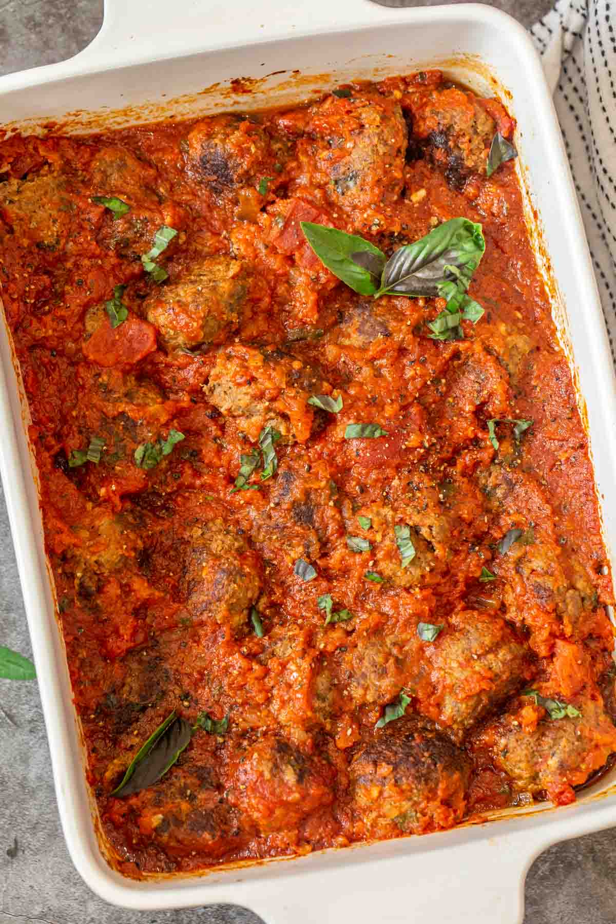 A white casserole full of juicy meatballs loaded with marinara sauce topped with fresh chopped basil. 