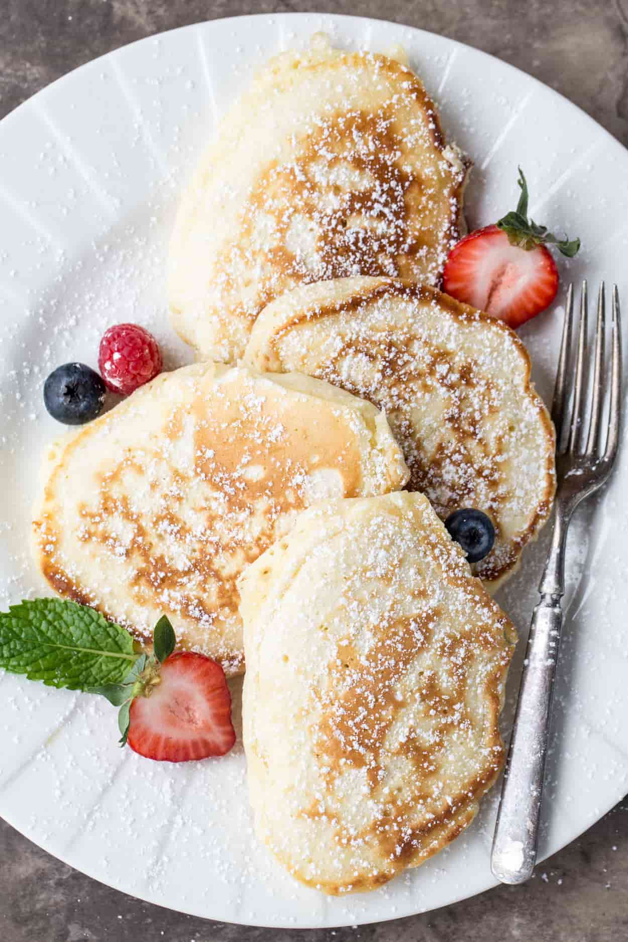 A white plate with four pancakes, sprinkled with powdered sugar. Beside the pancakes are fresh berries. 