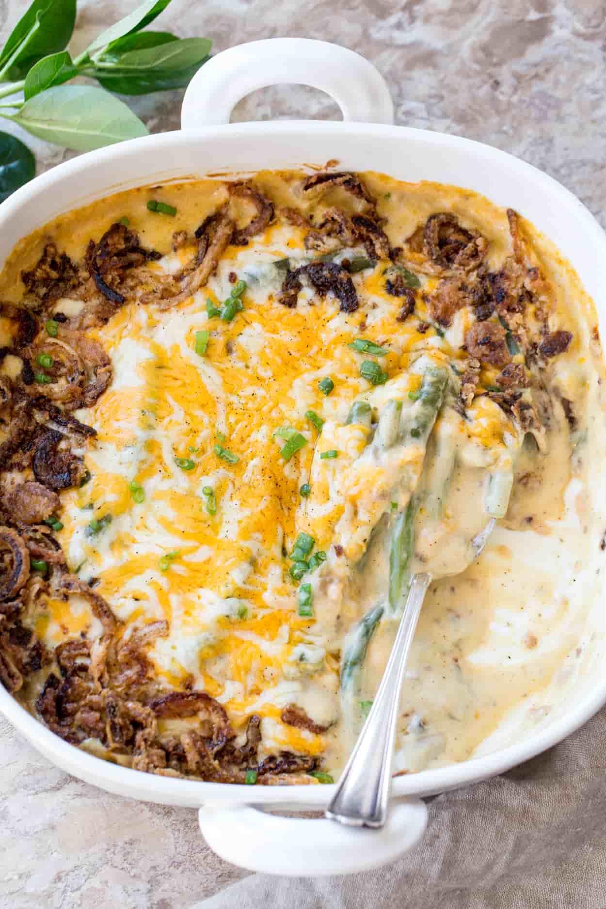 Green bean casserole in a casserole dish with a spoon. Topped with fresh greens, cheese, and fried onion rings.