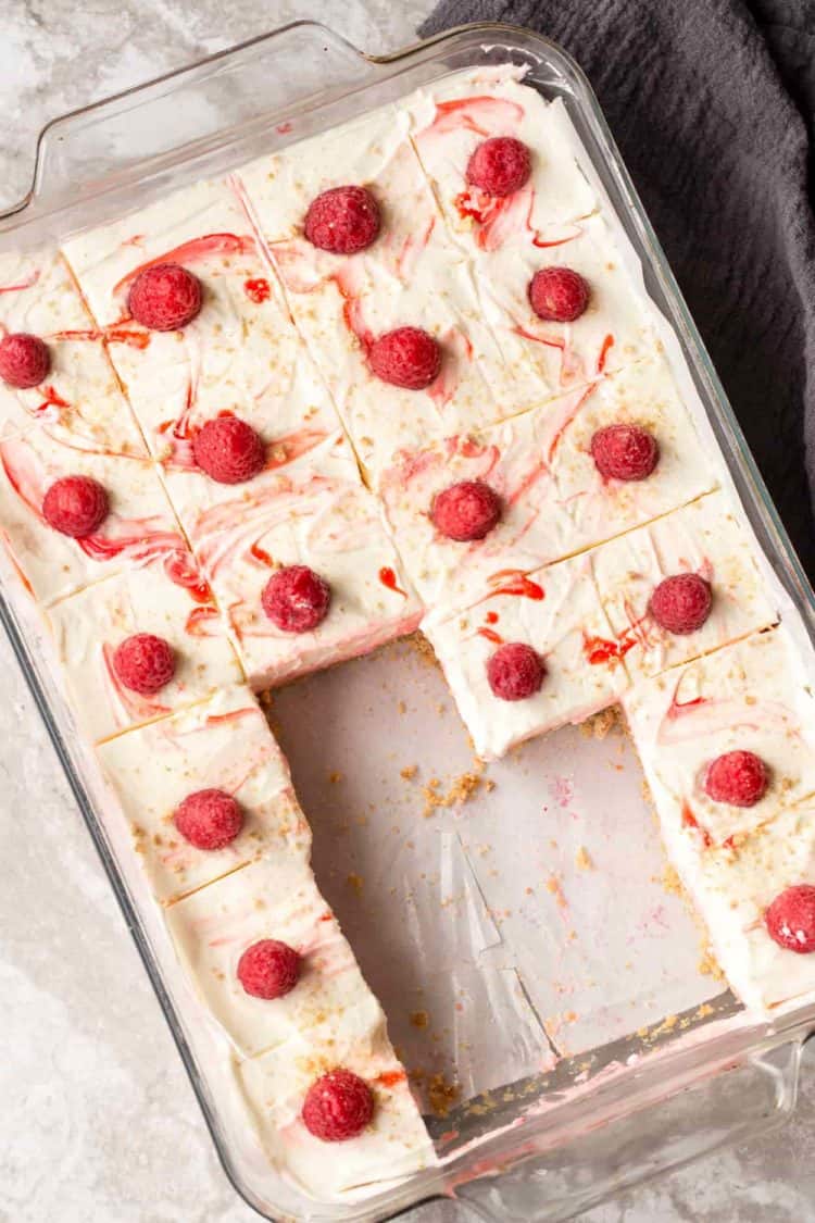Layered raspberry no bake cheesecake bars in a casserole dish. Bars cut out with raspberries on top.