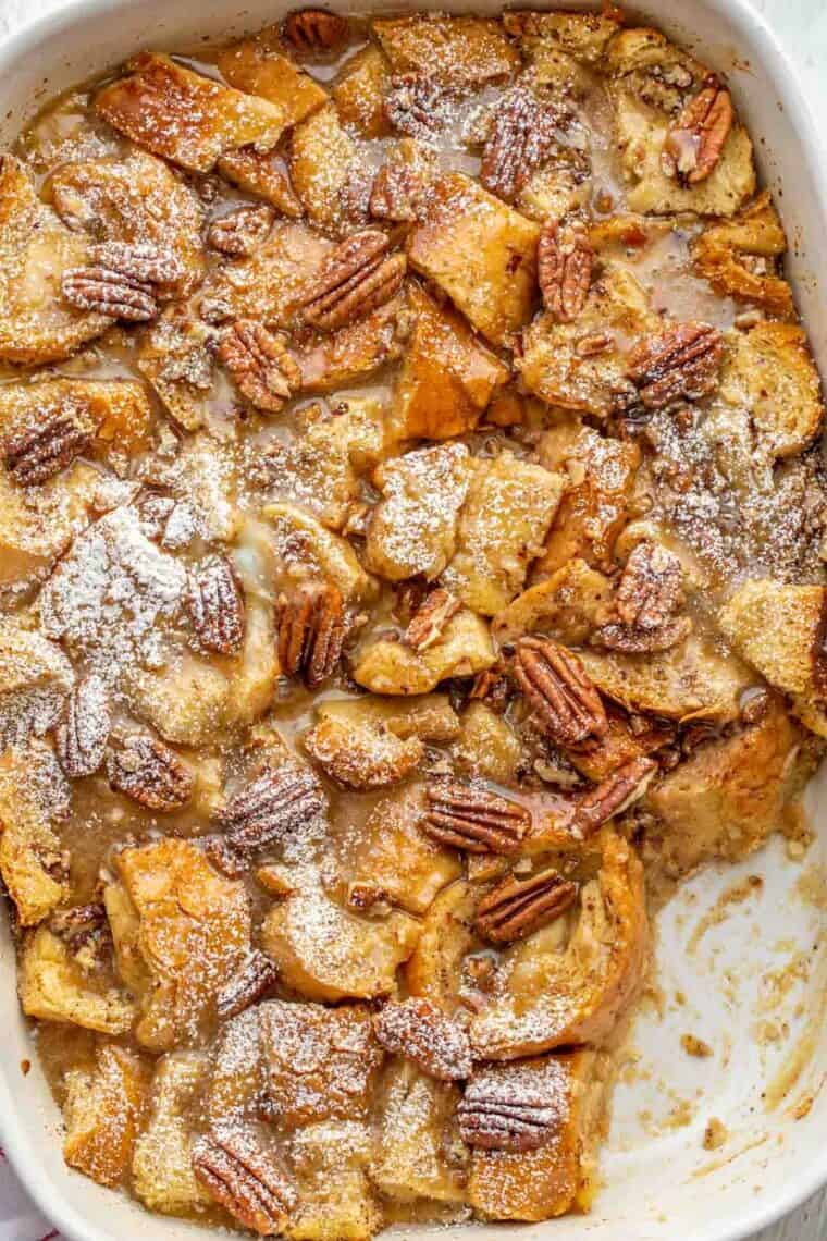 Baked maple glaze French toast casserole in a white baking dish topped with pecans and powdered sugar. 