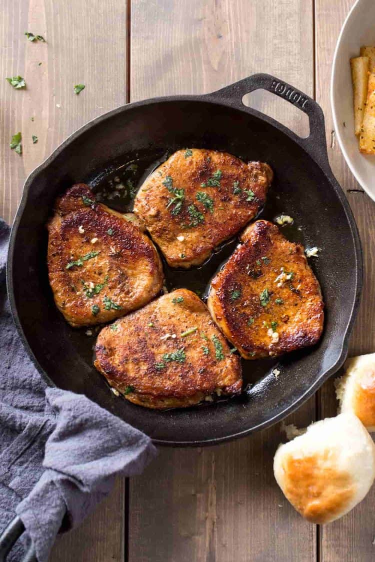 Pork chops in a skillet topped with garlic and greens. Next to dinner rolls.