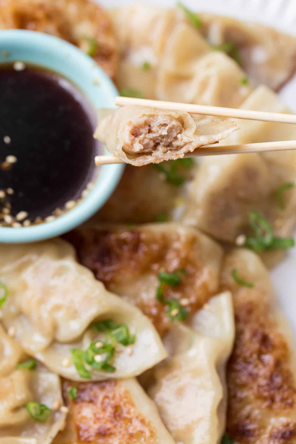Biten into dumpling in chopsticks next to a bowl of gyozas with dipping sauce. 