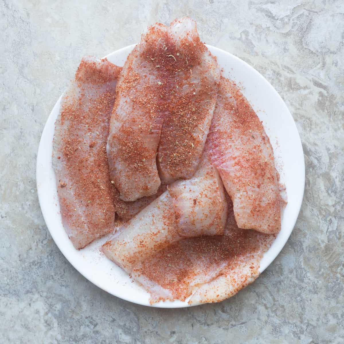 Seasoned tilapia stacked on each other in a white bowl.