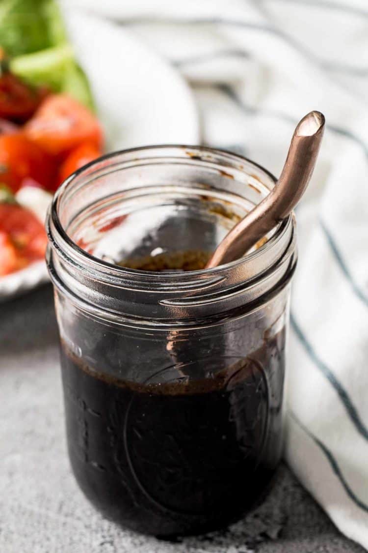 Balsamic Vinaigrette Dressing in a mason jar with a spoon, next to a bowl of caprese salad.