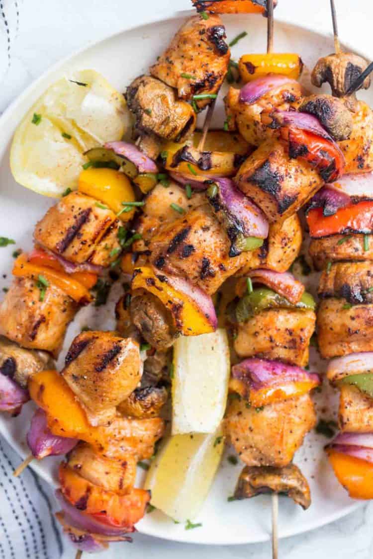 Grilled chicken kabobs on a white plate topped with greens and next to sliced lemons.