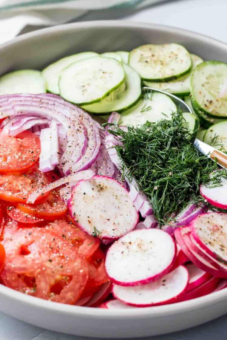 Sliced radishes, cucmbers, tomatoes, and onions in a bowl with a spoon and fresh chopped dill.