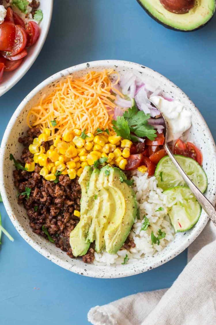 Simple taco bowl recipe in a bowl topped with corn, cheese, avocado and tomatoes.
