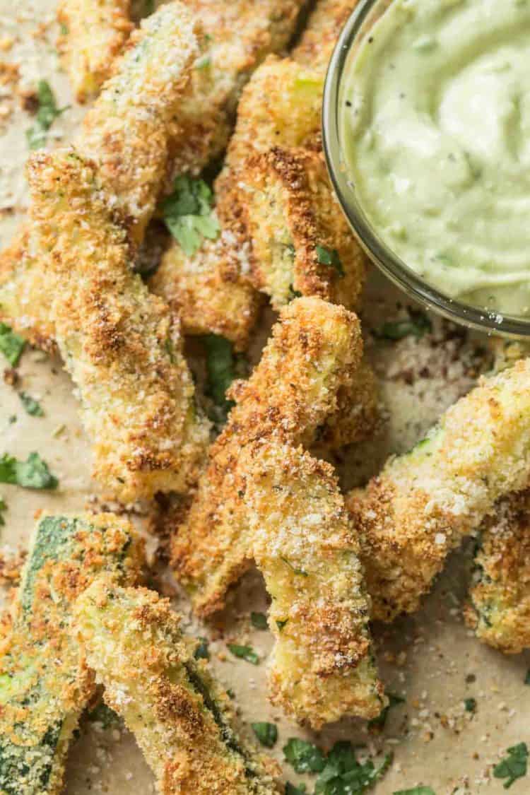 Oven baked zucchini fries on parchment paper next to a bowl of homemade avocado dipping sauce. 
