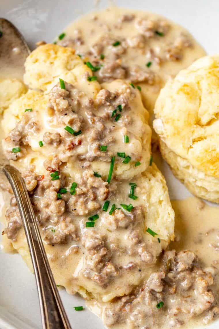 Homemade biscuits stacked on a plate loaded with sausage gravy and a spoon.
