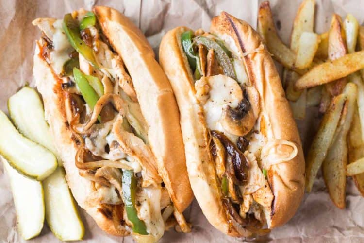 Two chicken philly subs next to french fries and sliced pickles.