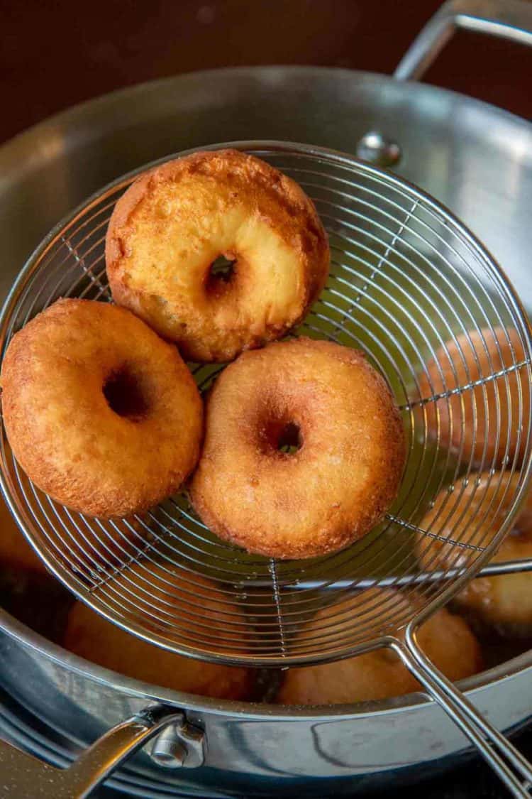 Three fried donuts in a strainer next to a frying pan with fried donuts. 