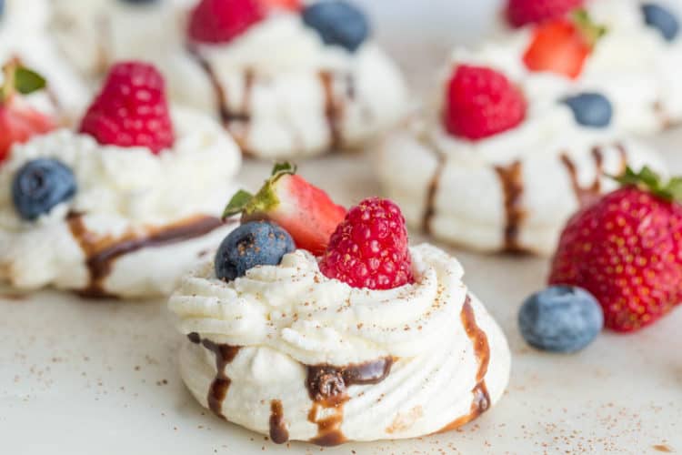 Meringue shell desserts with whipped cream on a platter topped with fresh berries and chocolate.