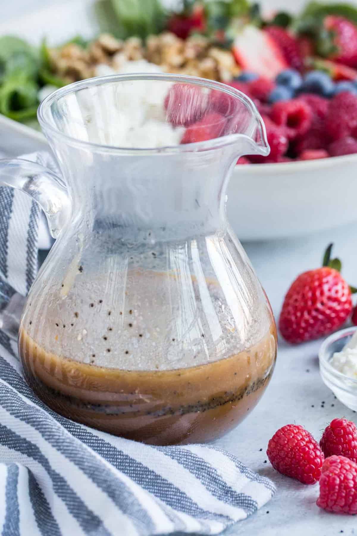 Homemade poppy seed dressing in a glass jar with a bowl of salad behind it.