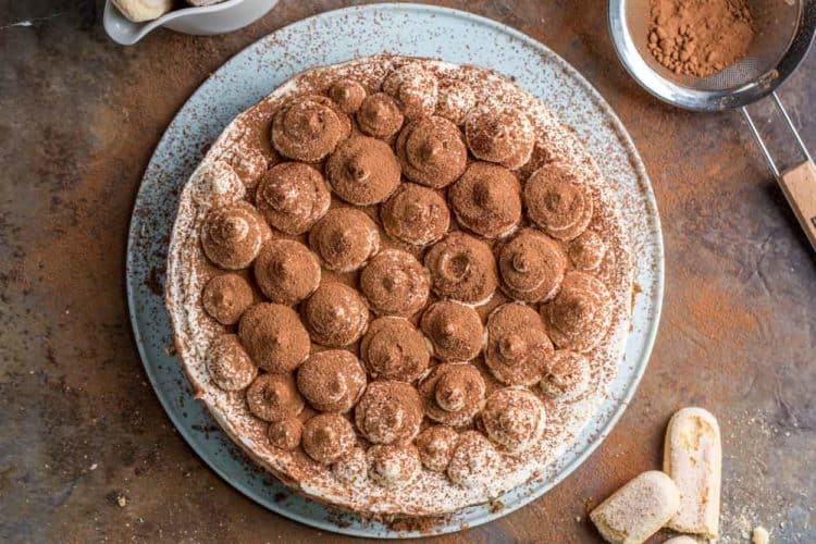 A whole Italian Tiramisu cake piped with frosting as decor and dusted with cocoa powder on a cake platter.