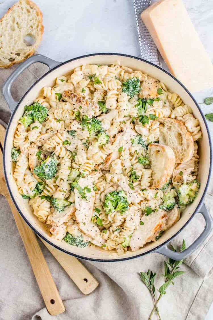 Chicken broccoli alfredo pasta in a pot next to spoons, bread and Parmesan cheese.