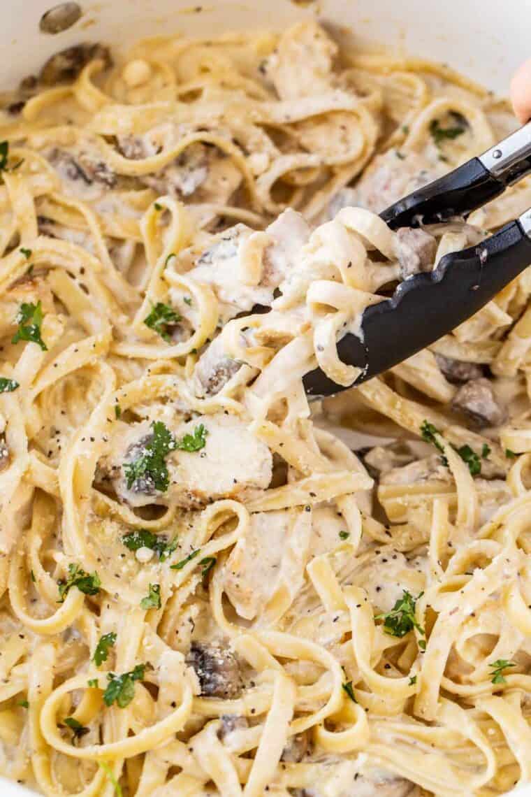Chicken fettuccine alfredo in a skillet with tongs topped with fresh greens and black pepper. 