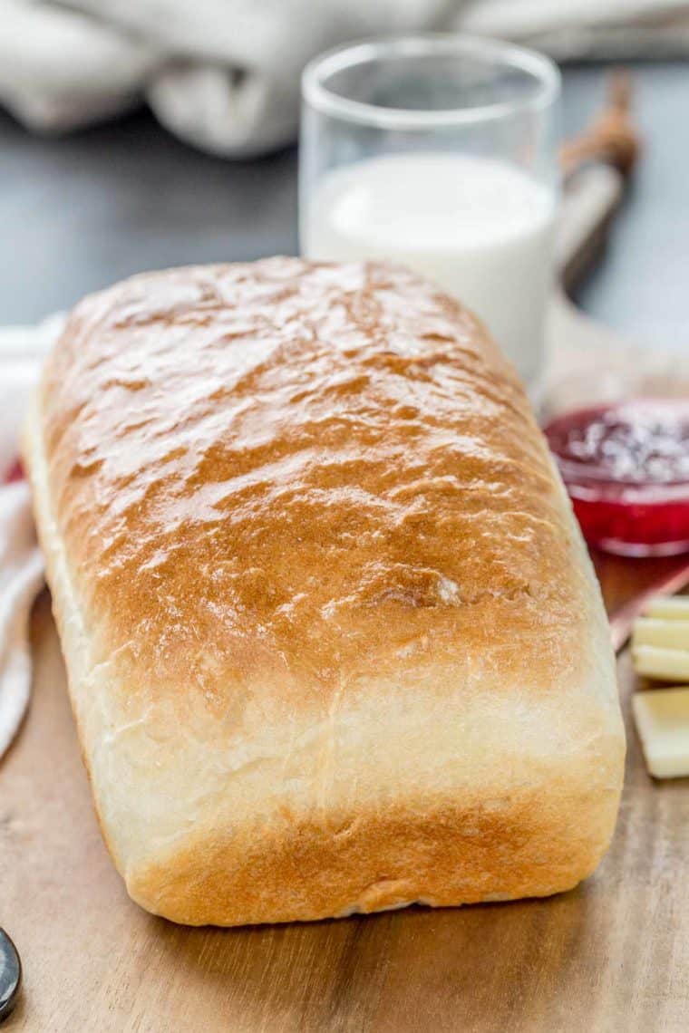A homemade bread loaf on a cutting board with a glass of milk, butter and jam.