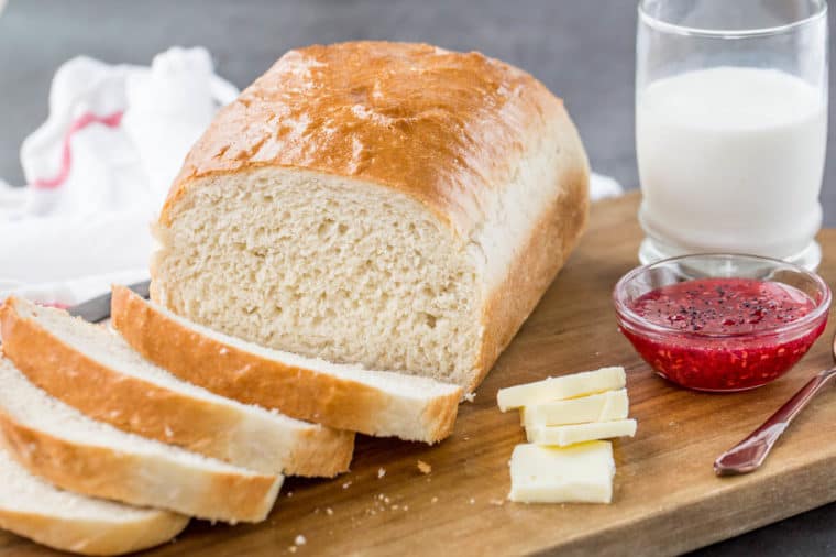 White bread loaf on a cutting board that is sliced next to butter slices, a glass of milk and a bowl of jam.