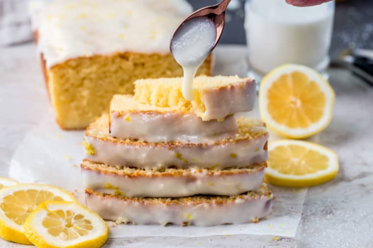Lemon bread load slices stacked on top of each next to lemon slices with a spoon of drizzling glaze.
