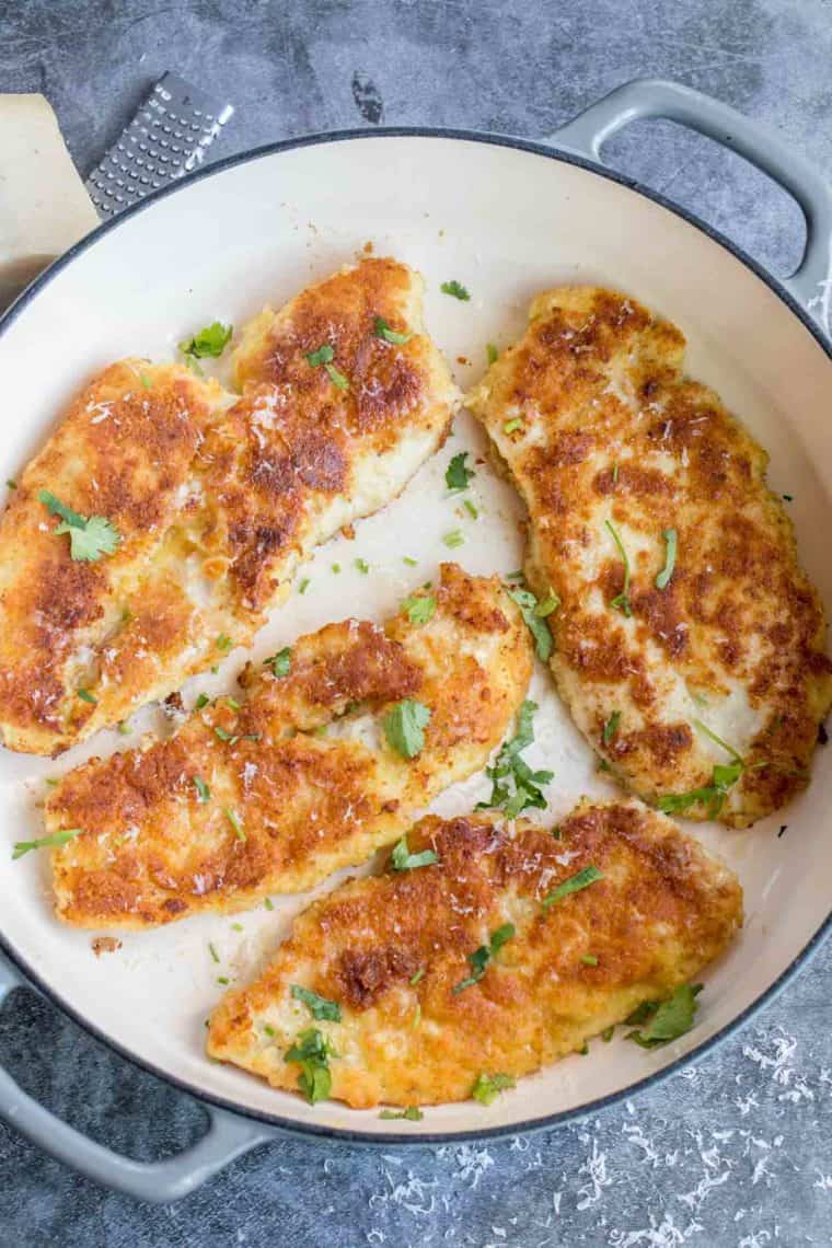 Four parmesan crusted chicken breasts in a skillet topped with fresh greens and grated parmesan cheese. 