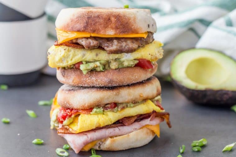 Breakfast sandwich on top of a breakfast sandwich around chopped greens, coffee mugs and an avocado. 
