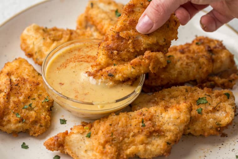 Chicken tenders on a plate with a chicken tender dipped into the dipping sauce. 