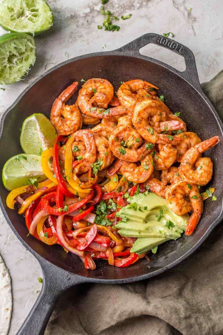 Shrimp, sauteed vegetables, avocados and limes in a black cast iron dish.