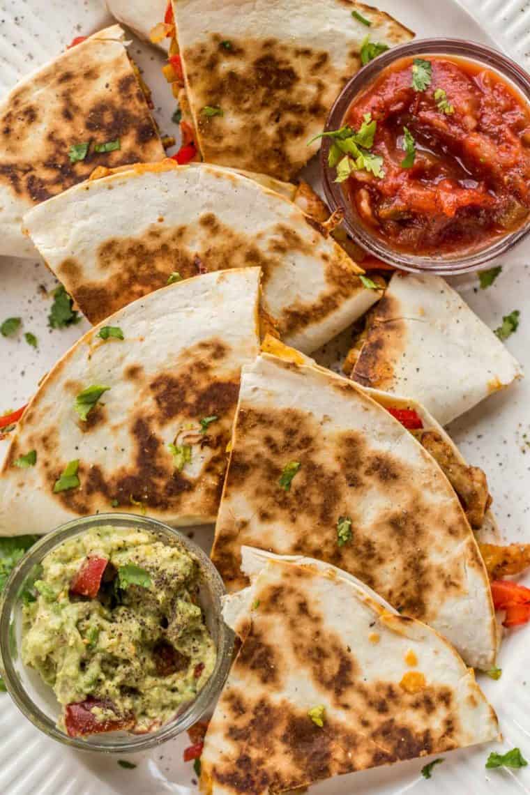 Quesadillas loaded with cheese, chicken and veggies laid out on a plate with a side of salsa and a side of guacamole topped with greens.