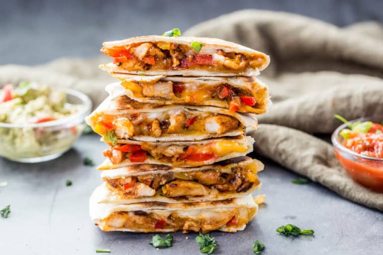 A stack of cheesy chicken quesadillas topped with fresh greens and next to a bowl of guacamole and salsa. 