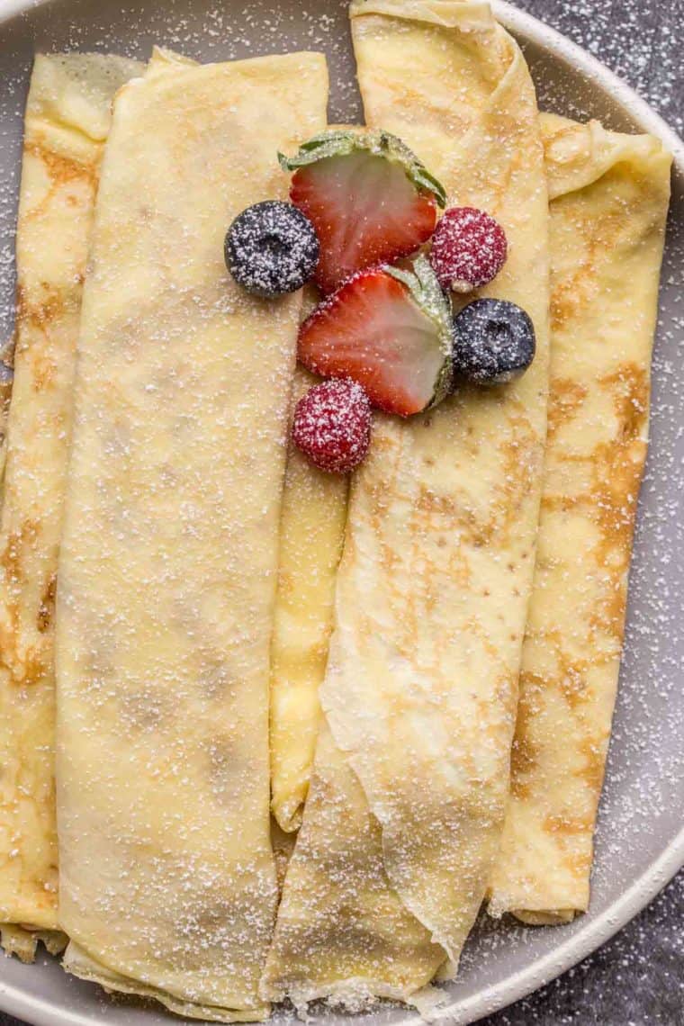 Homemade crepes on a plate topped with powered sugar and fresh berries. 