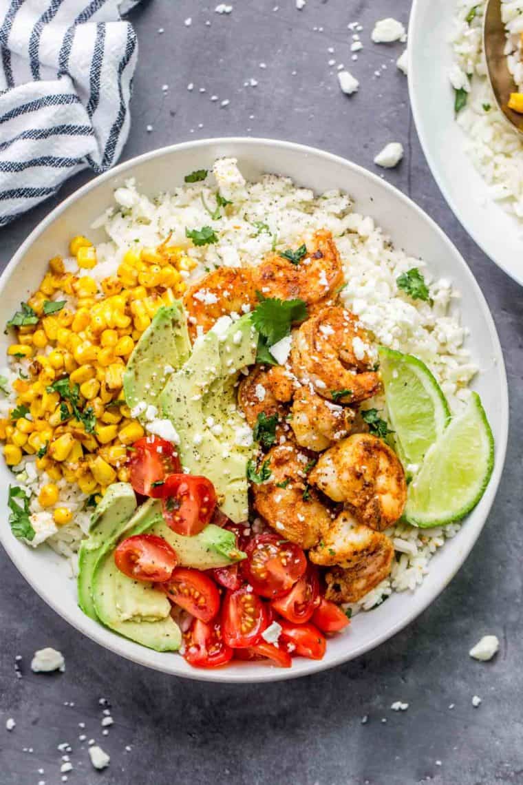 Rice bowl loaded with shrimp, corn, tomatoes and avocado topped with feta cheese next to a rag.